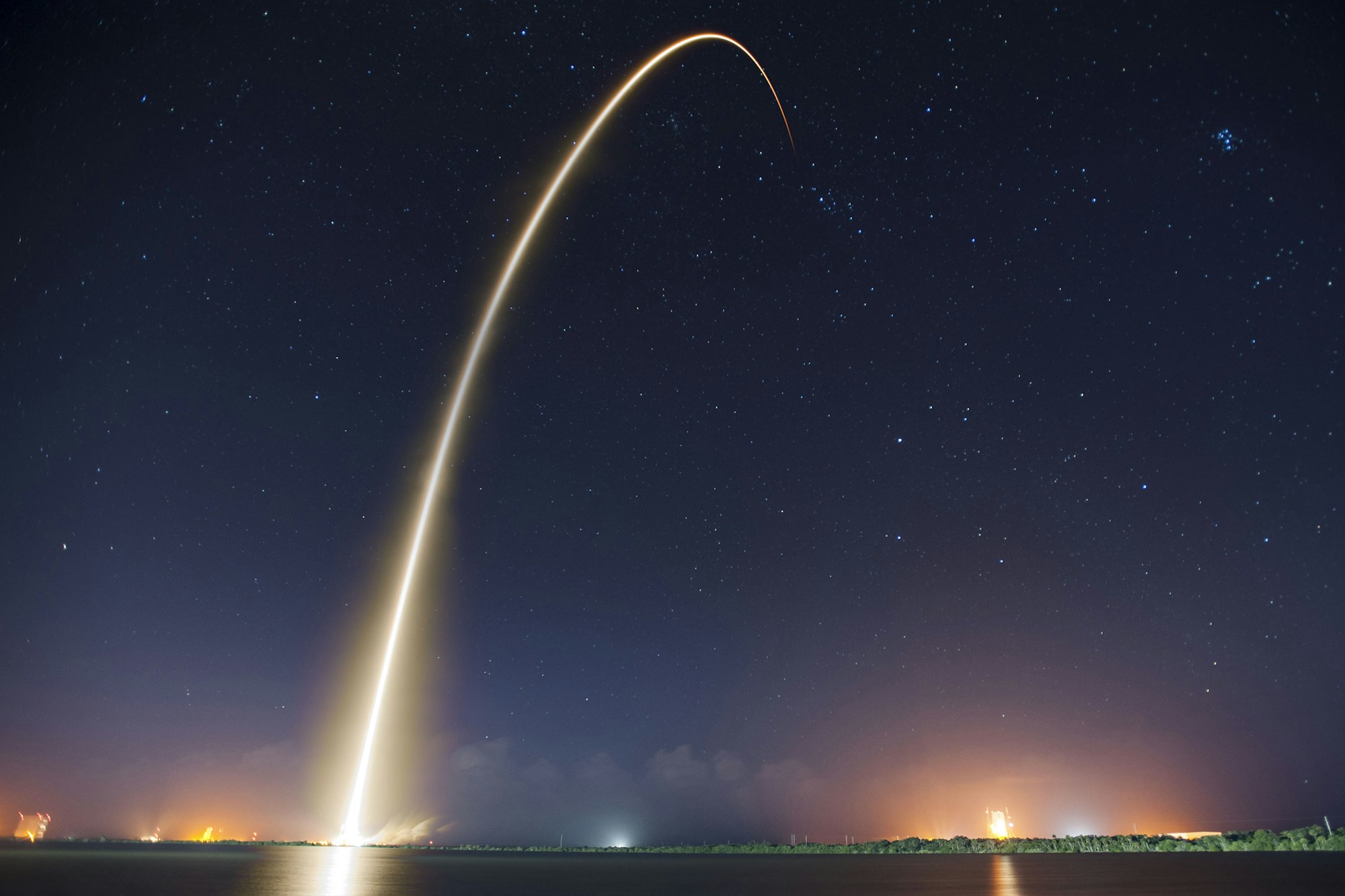 Rocket launching into a starry night sky, light trail arcing upward against a dark purple horizon
